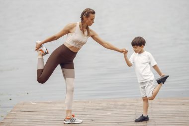 A mother and son holding a yoga pose together on a wooden pier by the water, promoting family fitness.
